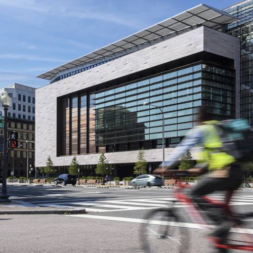 An exterior shot of a towering building facade, with a cyclist racing by in the foreground.