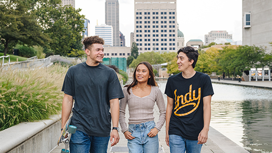 Two men and one woman walking beside a river with Indianapolis in the background.