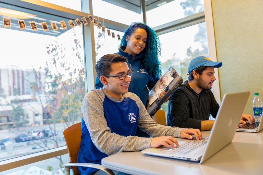 female Residential Life staff in duty uniform standing and smiling behind two male students sitting at a table with laptops open