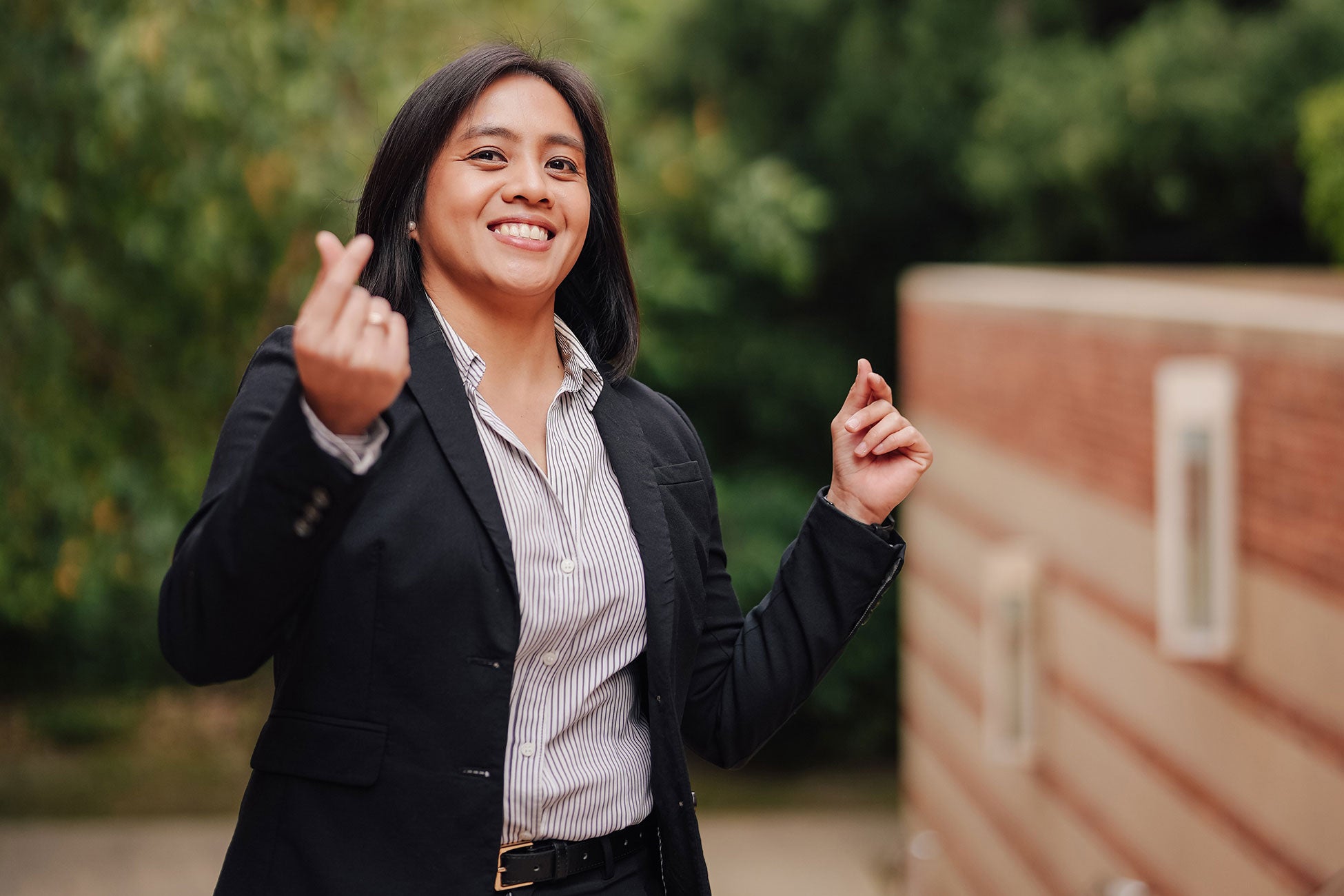 A dark-haired woman in business attire stands outdoors and smiles to camera while snapping fingers