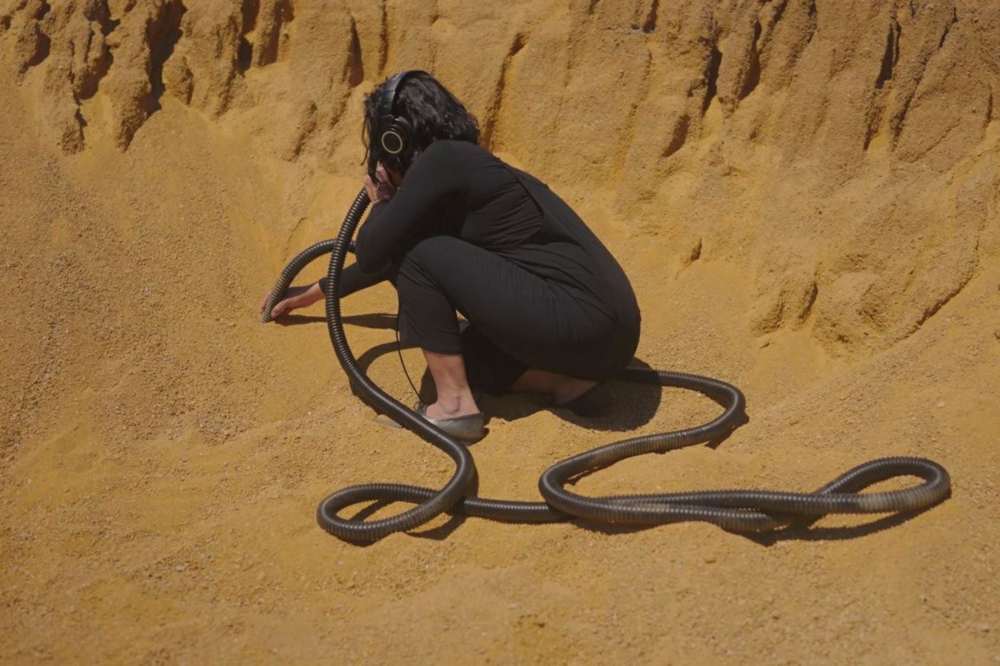 A person kneeling down on sand with headphones, listening to an installation through a tube.