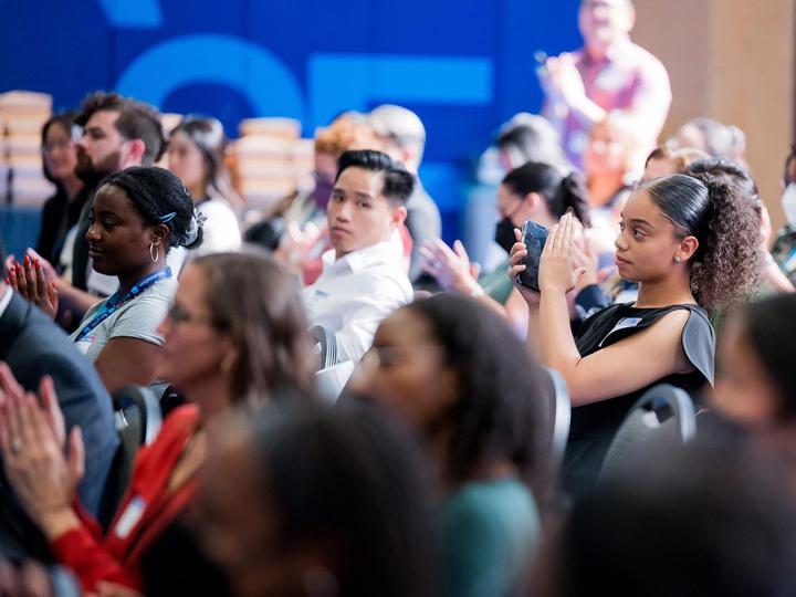 Participants of the 2023 HEAR Symposium are seated in a well-lit gymnasium clapping and watching a speaker at the front of the room out of view.