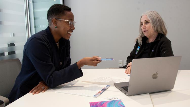 UCSF CARE Advocates Kendra Hypolite, on the left and Denise Caramagno on the right are seated inside a conference room. Kendra is pointing to a laptop in mid conversation with Denise seated across the table.