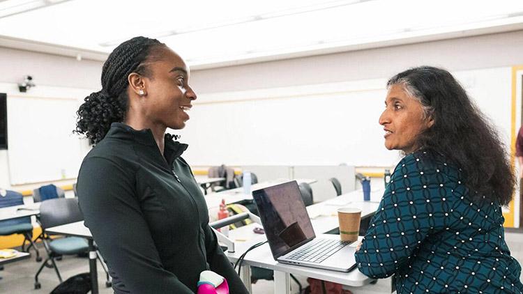 Two women having a conversation at the front desk in an empty classroom.