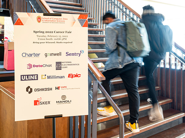 Two students walk up stairs next to a Career Fair sign listing a dozen employers.