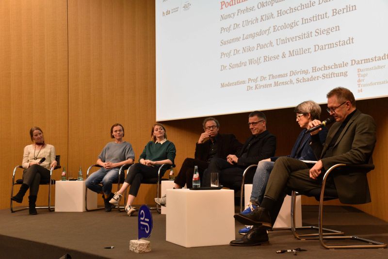 A panel discussion with seven people sitting in front of an audience. In the background is a large screen with the names of the speakers and moderators.