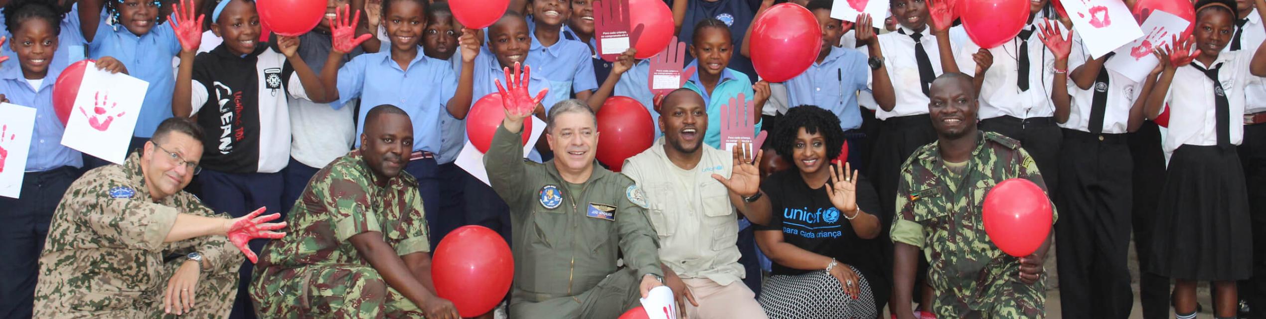 A group of children with training mission soldiers smile and wave.