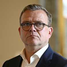 Close-up of a man with round horn-rimmed glasses and a serious expression, wearing an open-necked shirt and dark blue blazer.  