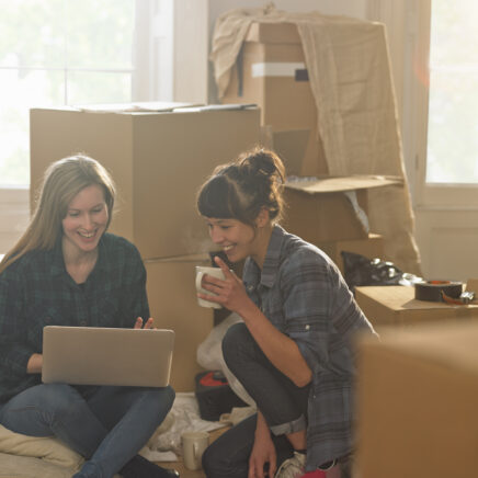 Deux jeunes femmes professionnelles assises dans un nouvel appartement entouré de boîtes d'emballage, elles boivent du thé et regardent un ordinateur portable.