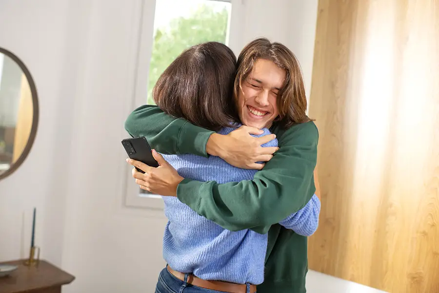 Ado enlaçant sa mère, il tient un smartphone dans sa main et sourit