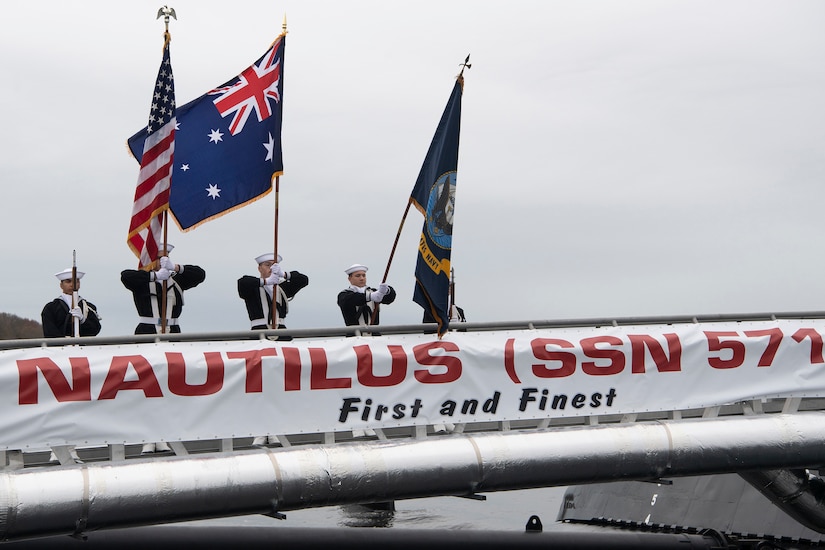 Three service members hold flags as they stand behind a sign saying "Nautilus SSN 571, First and Finest."