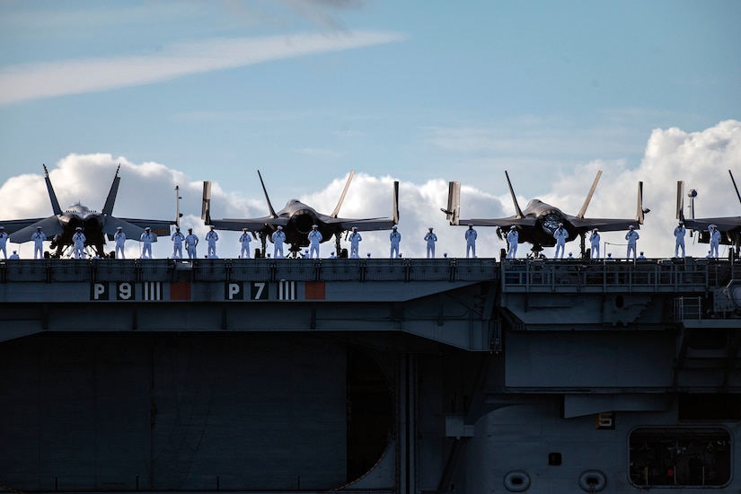 Four parked fighter jets rest behind a line of sailors on the deck of an aircraft carrier in a clear day.