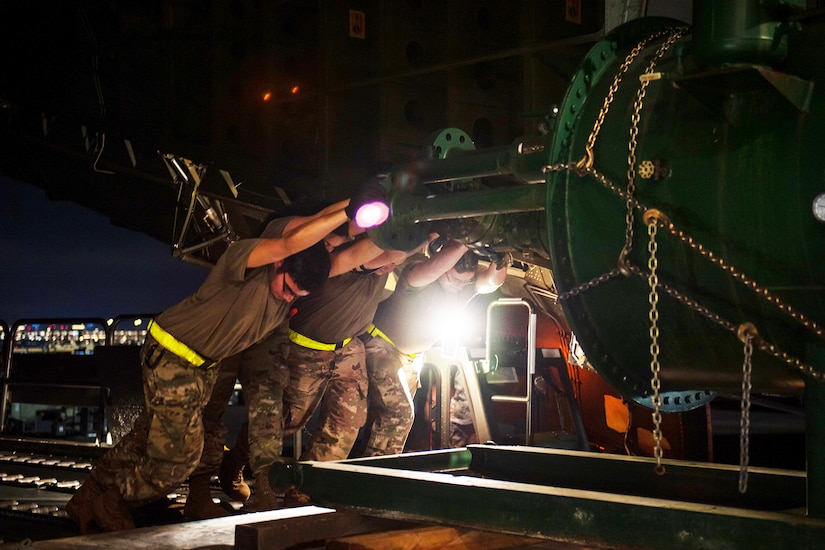 Three airmen push heavy equipment onto an aircraft loading bay at night.