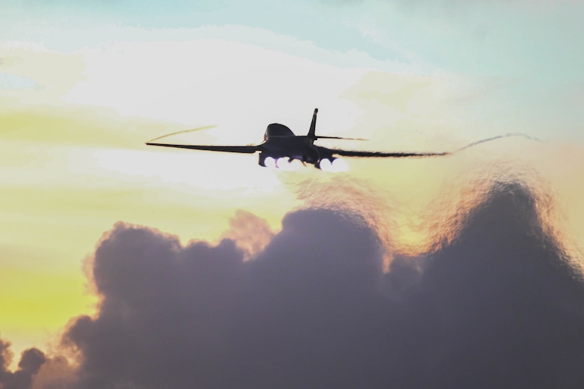 The back of a large aircraft is seen as it takes off into a yellow and light blue sky above billows of grey smoke.