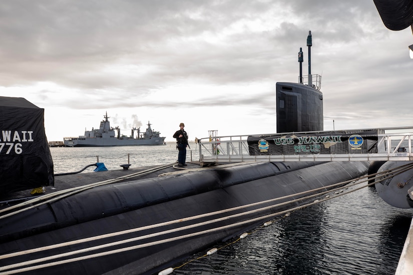 A sailor stands on top of a submarine next to a gangplank with a “USS Hawaii” banner along the side on an overcast day. Another military ship floats in the background.