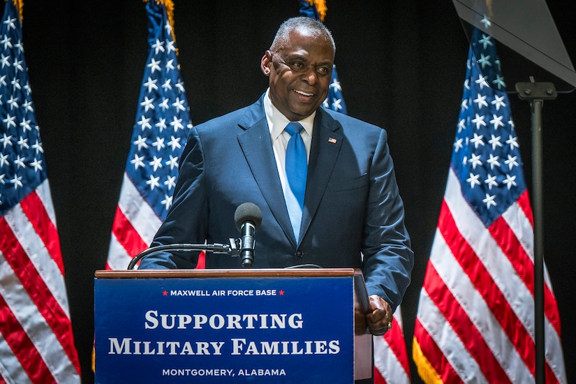 Secretary of Defense Lloyd J. Austin III smiles at a lectern with a sign that reads "SUPPORTING MILITARY FAMILIES," in front of U.S. flags.