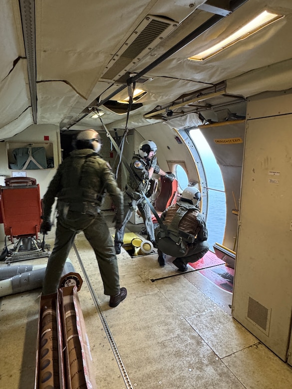 Three sailors stand next to an open doorway in an airplane cabin and look out at sea.