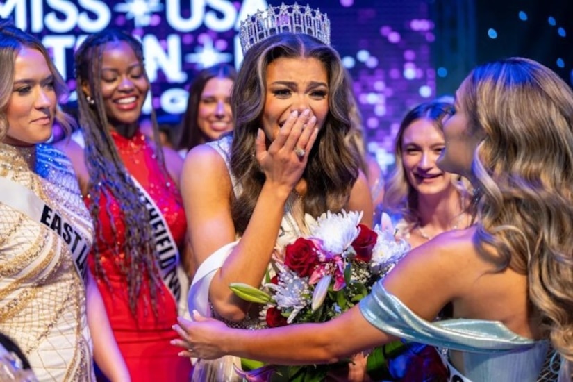 A woman wearing a crown, Miss USA sash and flowers is congratulated by other contestants as she cups her hand over her mouth.