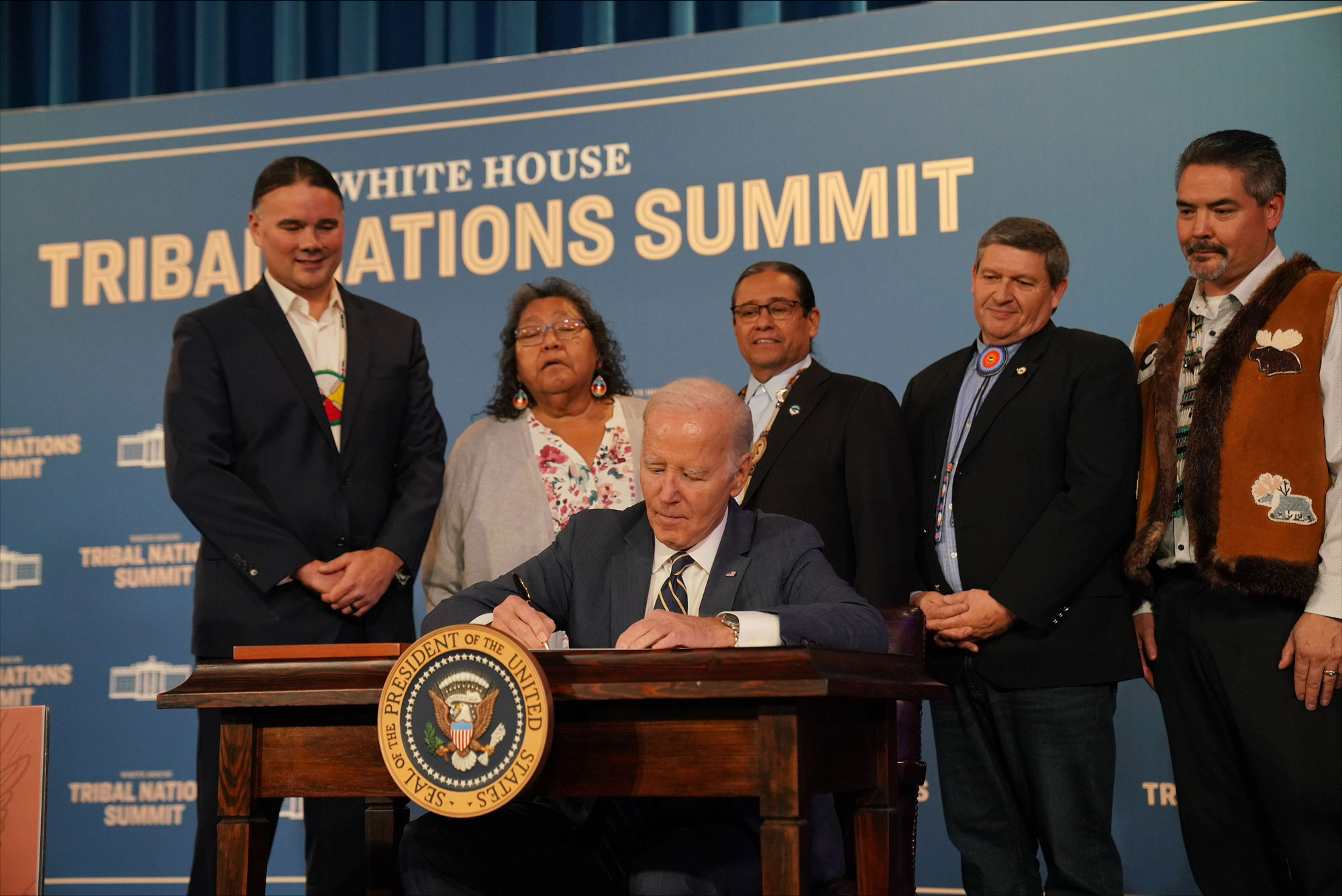 President Biden signing paper at table with 5 others standing behind him