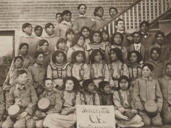 Archive photograph of a group of Indigenous children posed outside a Federal Indian Boarding School. 