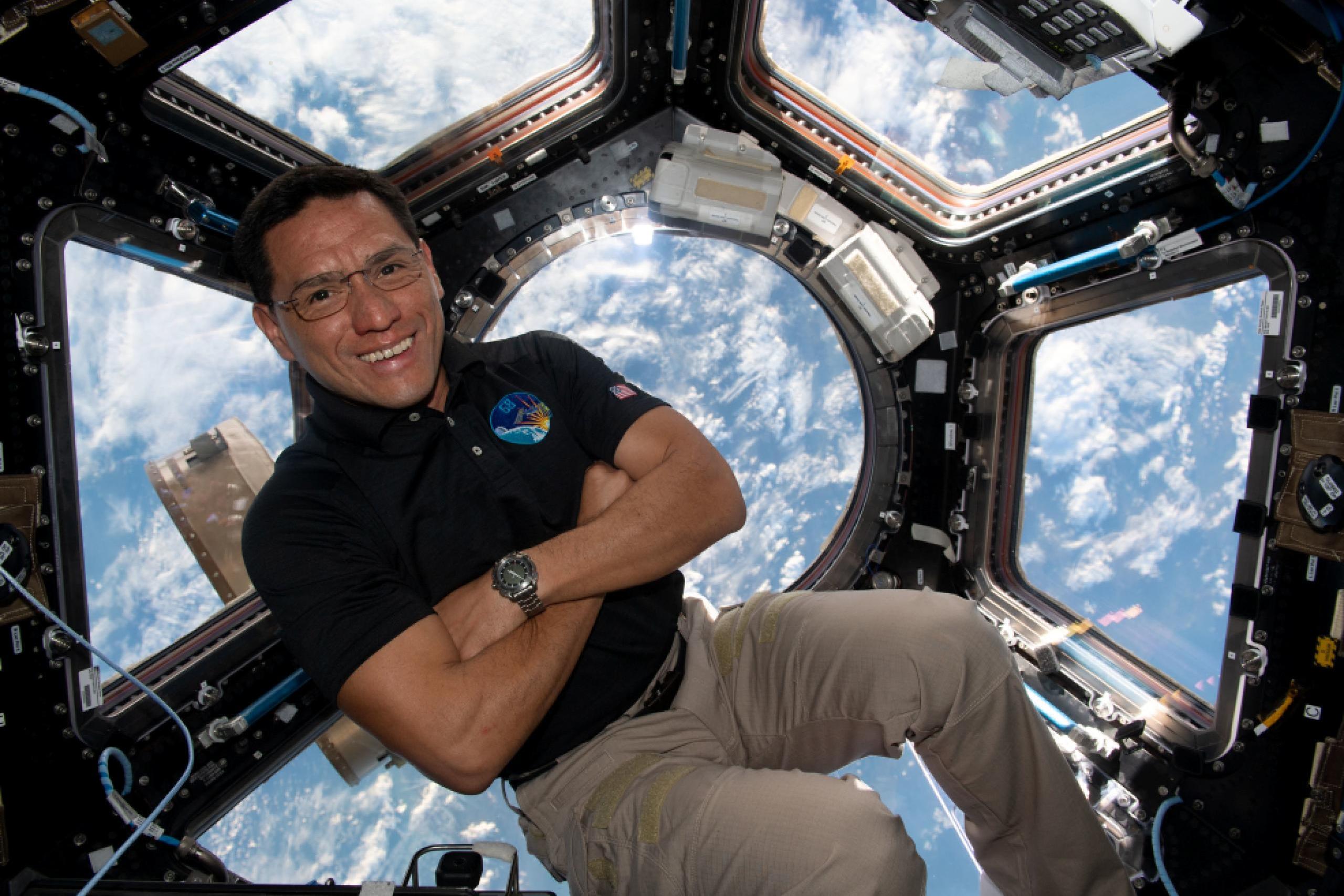 Imagen de Frank Rubio flotando en la cúpula de observación de la Estación Espacial Internacional. Rubio, un hombre de pelo y ojos oscuros y con lentes, sonríe y tiene los brazos cruzados. Va vestido con un polo oscuro y pantalones khaki. Detrás suyo se ve la atmósfera de la Tierra a través de las ventanas de observación de la cúpula.