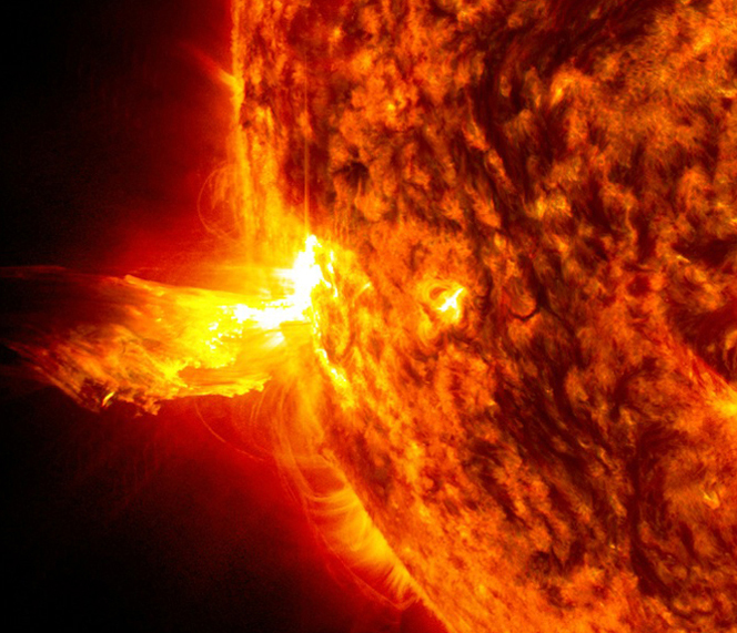 A portion of the Sun, appears in red, yellow, orange, and black. A yellow burst of solar material juts off of the Sun, against the black expanse of space.