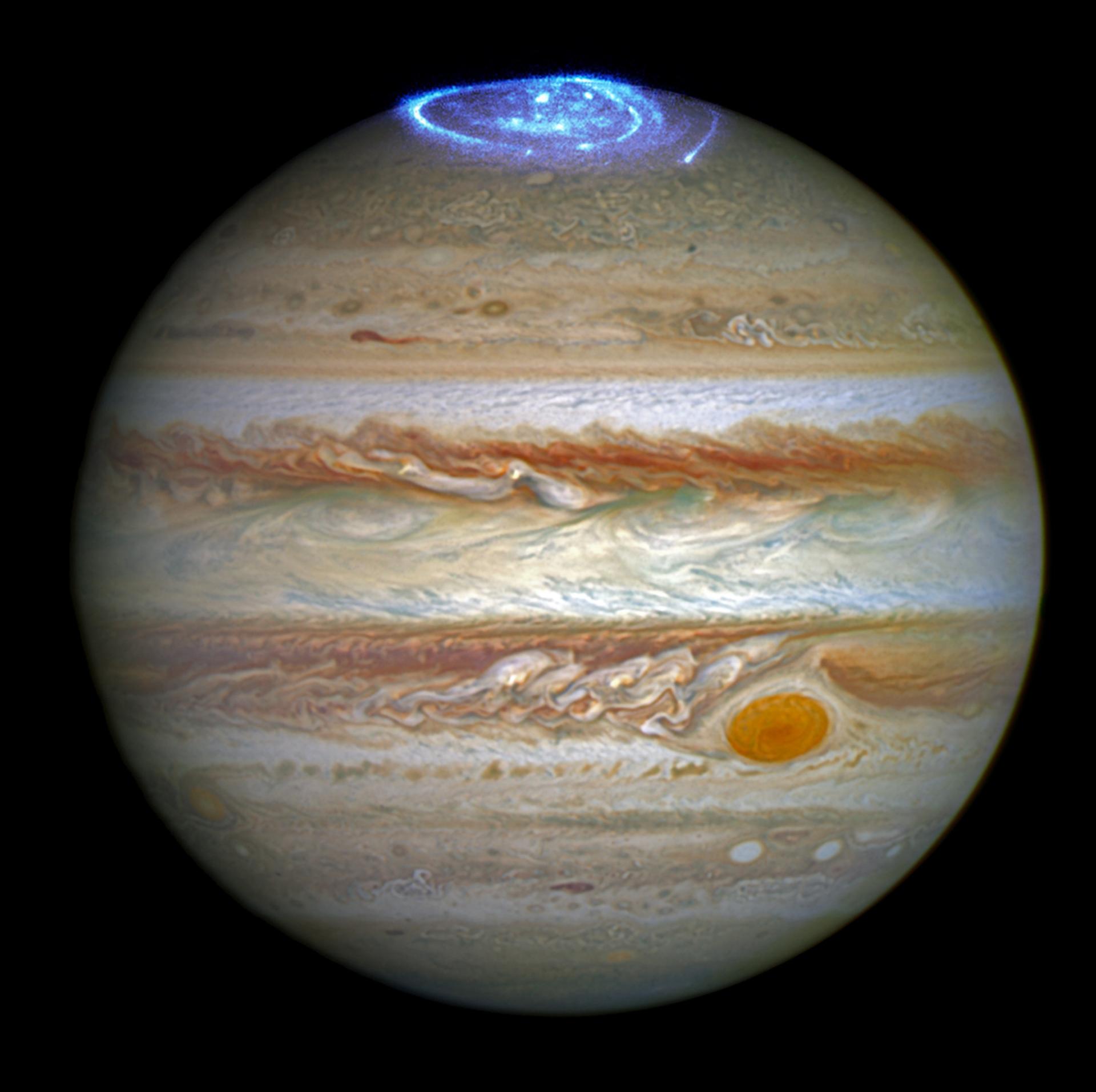 The planet Jupiter is seen against black space. At the planet's northern pole, its aurorae are seen as a bright blue whirl of light.