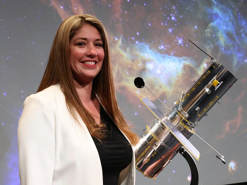 Headshot of Lauren Miller standing in front of a 3D model of the Hubble Telescope.