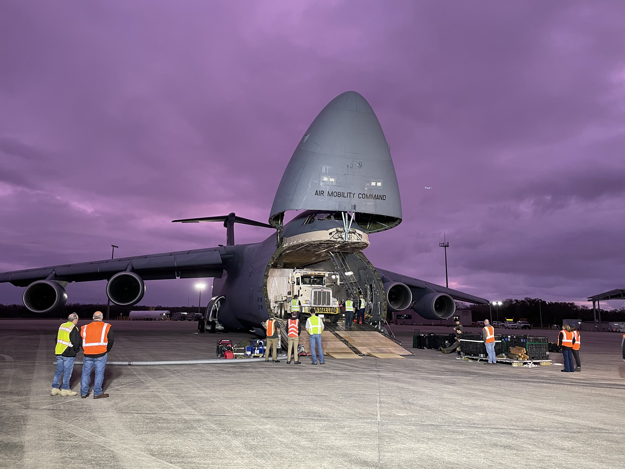 A large cargo plane sits on the runway with the nose cone open to reveal a semi truck inside unloading the GOES U satellite at KSC.