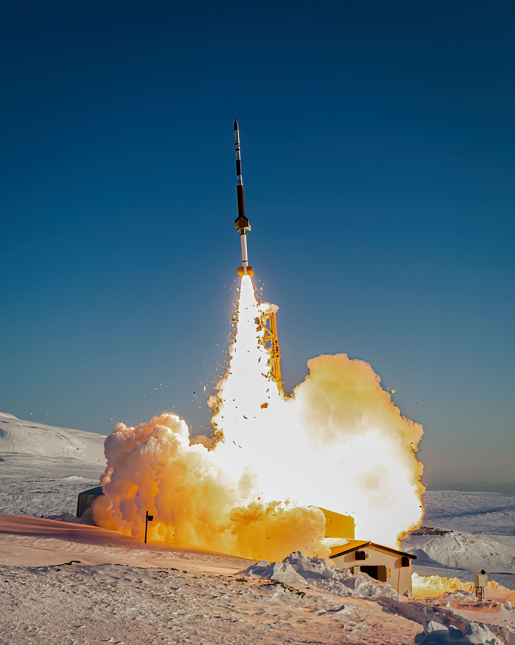 A rocket launches into the blue sky from a snow-covered launch range, leaving a bright cloud of rocket exhaust in its wake.