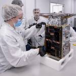 An engineer in a white lab suit holding a tape ruler to a small, microwave-sized, rectangular small satellite. One side has black panels, and others have silver panels and foil. Three team members are seen in the back.