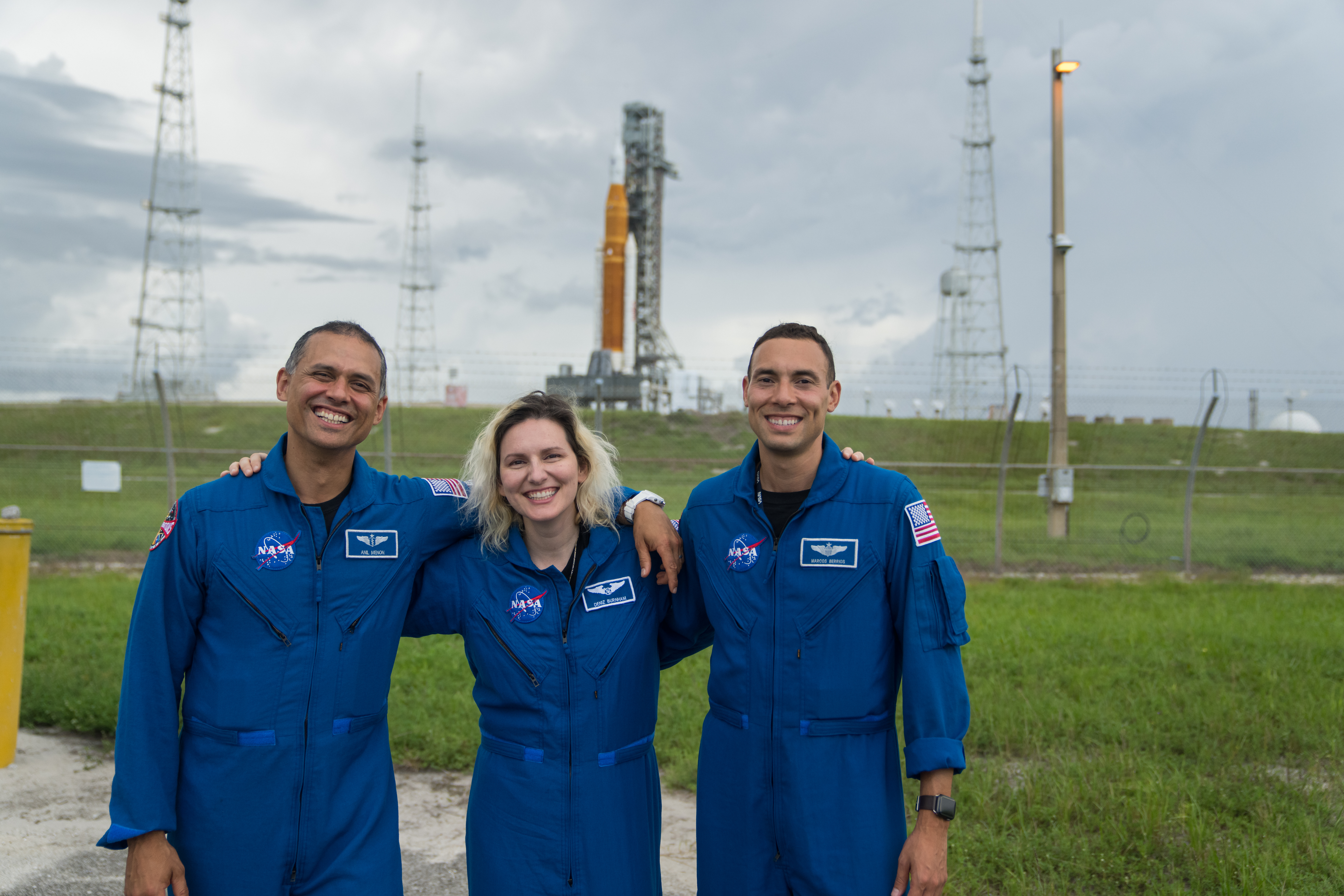 Three astronauts have their arms over each other's shoulders as they smile for the camera. From left to right, they are Anil Menon, a Ukrainian and Indian American man, Deniz Burnham, a white woman, and Marcos Berríos, a Latino man. All three wear blue jumpsuits with various patches sewn on, including a NASA patch, a patch with their name, and an American flag patch. The picture shows them from the knees up. In the distance behind them, beyond a grassy area, are the Artemis I rocket and spacecraft. The launch pad is out of focus, but the SLS's distinctive butterscotch orange color can be made out.