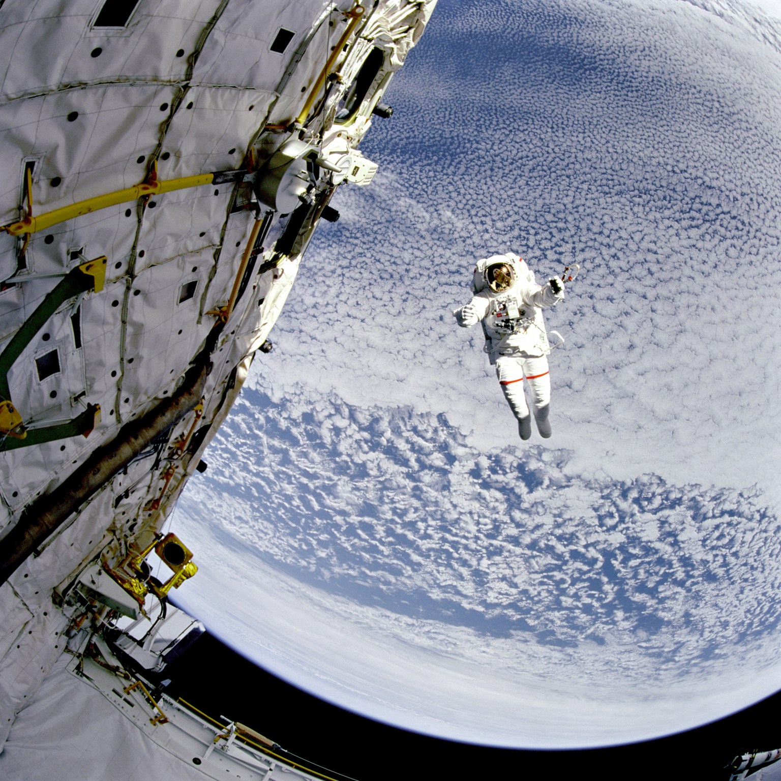 An astronaut in a white spacesuit floats untethered in space, next to a spacecraft. White clouds on Earth form a backdrop to this image.