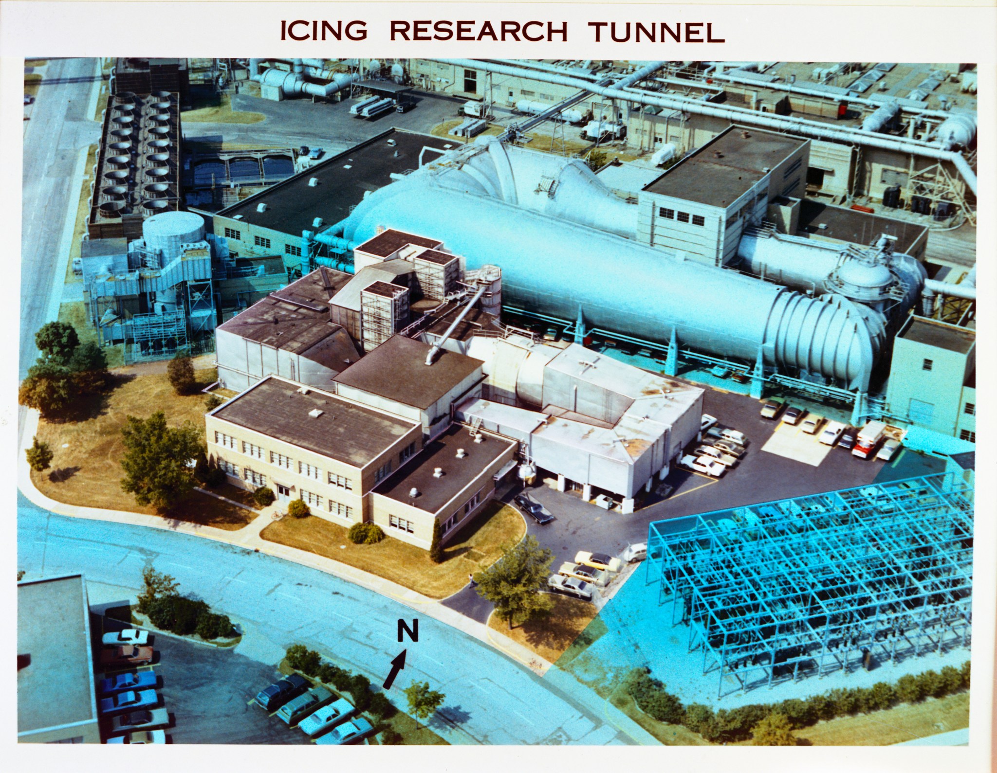 An aerial view of the Icing Research Tunnel. A cluster of building can be seen beside a large wind tunnel along with cars and road areas. A title at the top of the photo says, “Icing Research Tunnel.”