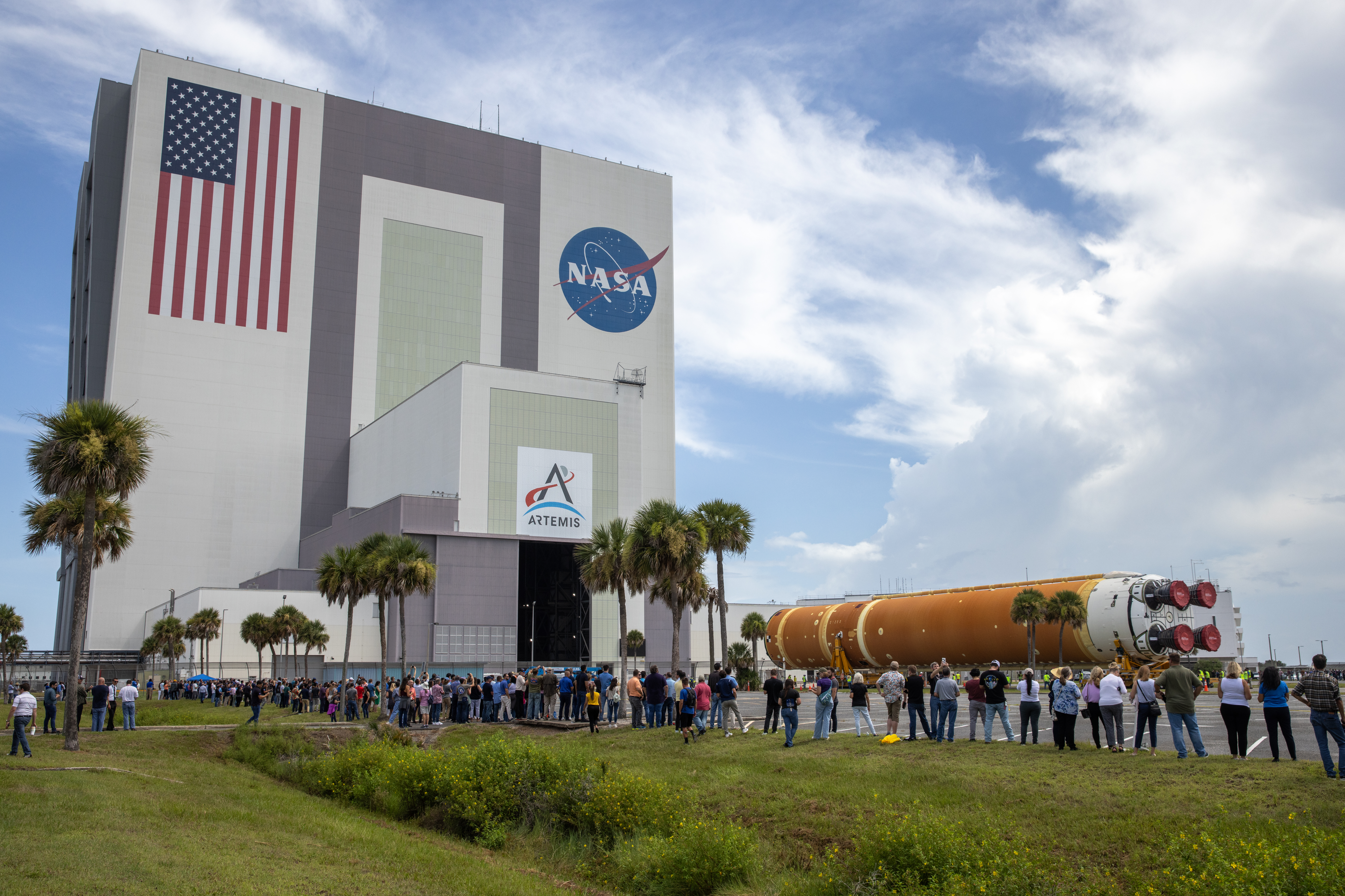 The Artemis II rocket core stage, a long orange cylinder with four red boosters) is on its side as it rolls into the Vehicle Assembly Building (VAB) at NASA's Kennedy Space Center while employees watch. The VAB is a tall rectangular building with the American flag and NASA "meatball" logo painted on its left and right side, respectively. There is also an Artemis logo on a lower portion of the building.