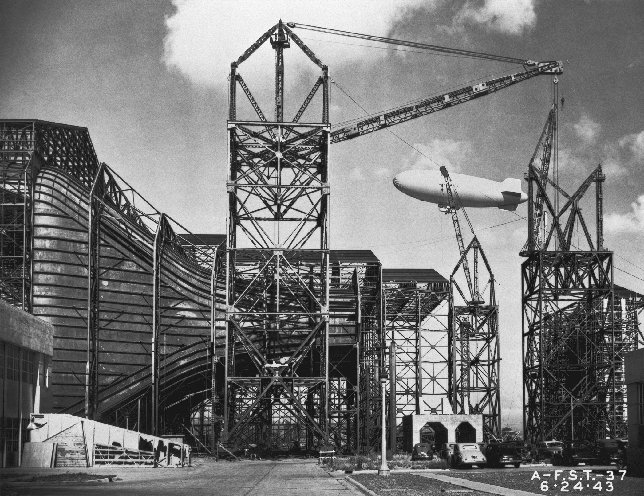 Black and white photo of a wind tunnel at Ames under construction. A Navy blimp is flying in the background.