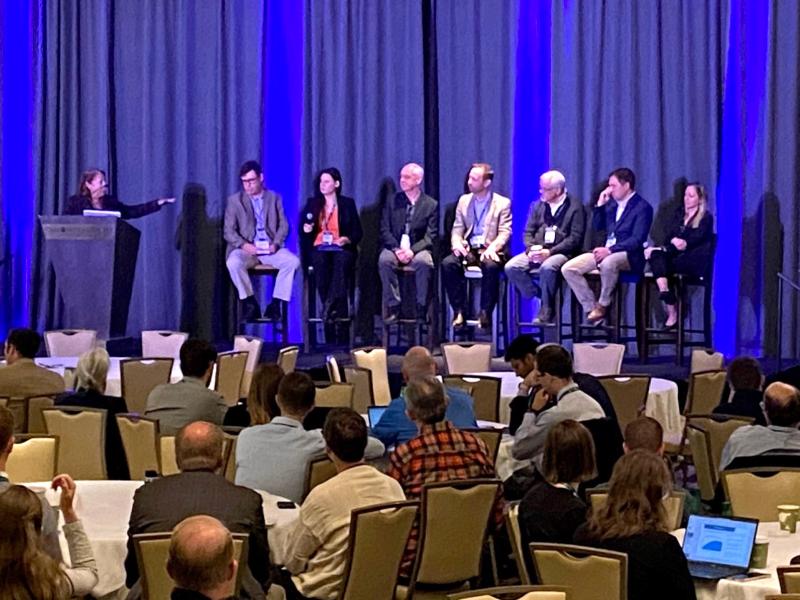 Seven speakers and a moderator on a stage at a conference.