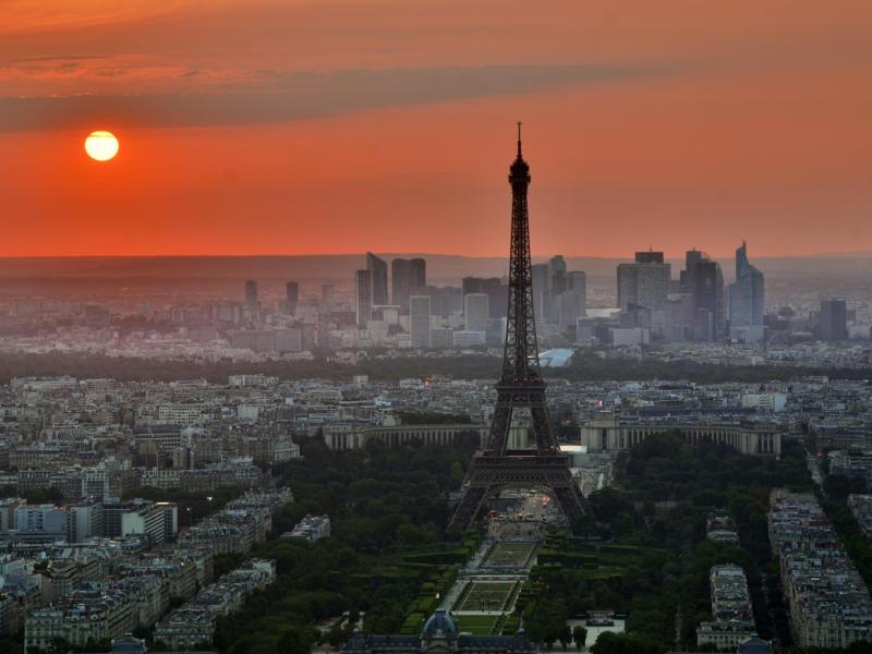 The glowing orange Sun shines over a lightly hazy Paris, the Eifel Tower standing erect, just right of center-frame.