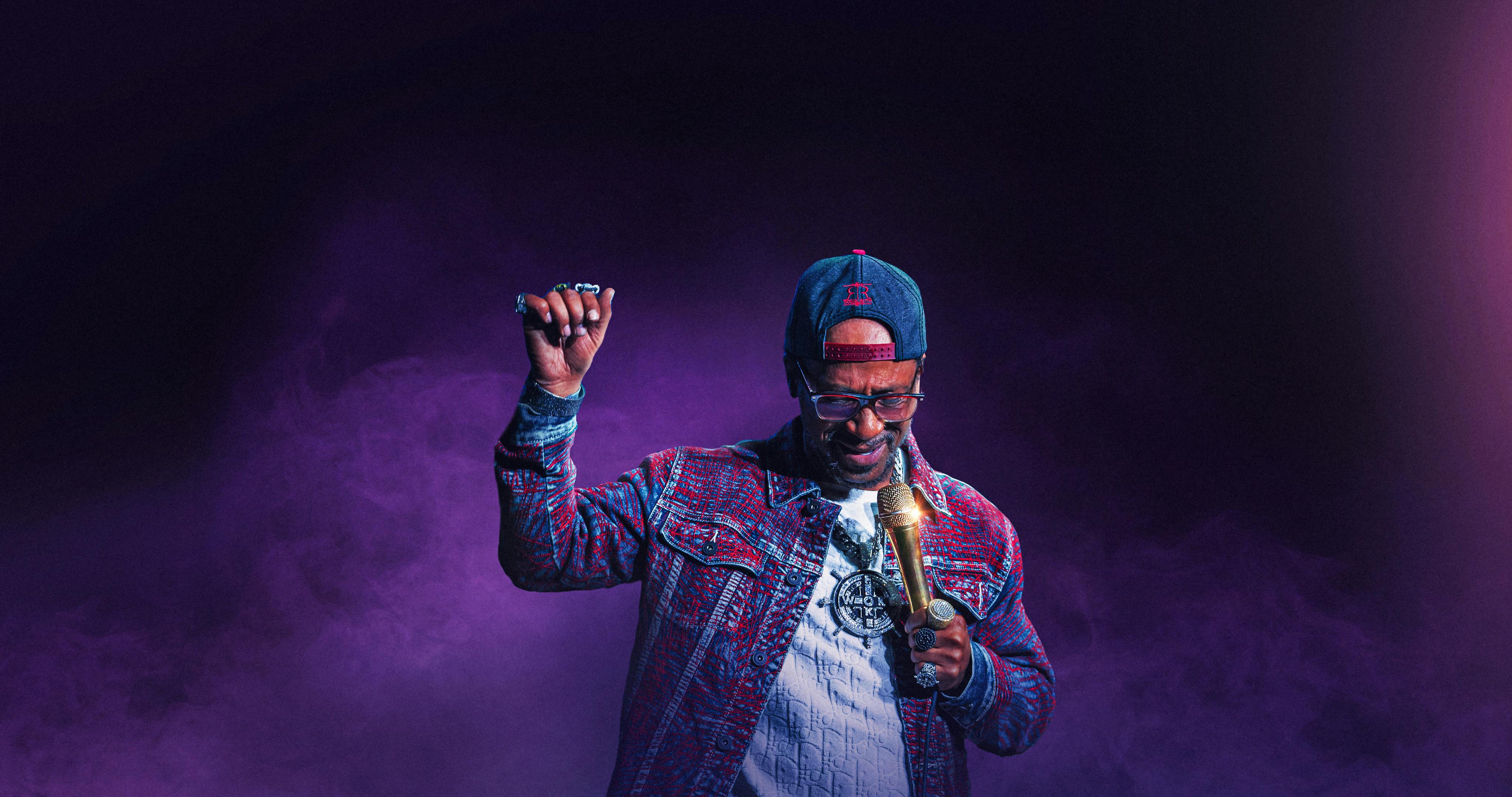 Katt Williams wears a beautiful patterned jacket against a foggy purple backdrop.