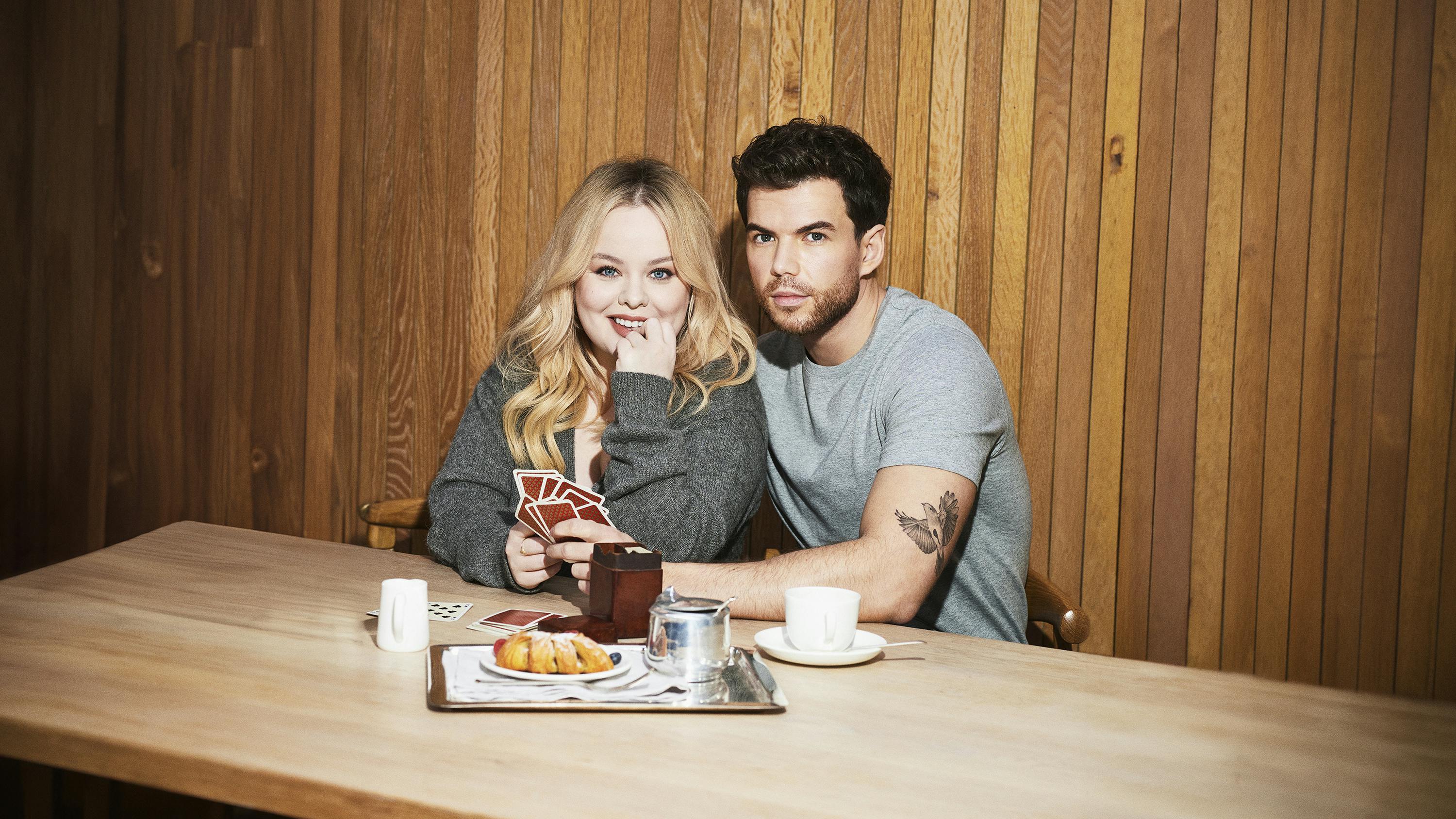 Nicola Coughlan and Luke Newton sit at a wooden table playing cards.