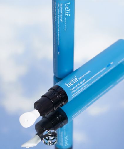 A tube of Belif Aqua Bomb eye cream is placed on a reflective surface next to its removed cap. The blue tube shows the product partially. A small amount of the cream is visible next to the tube.