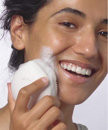 A smiling woman is cleaning her face with the LG Pra.L Wash POP Ultrasonic Facial Cleansing Brush.