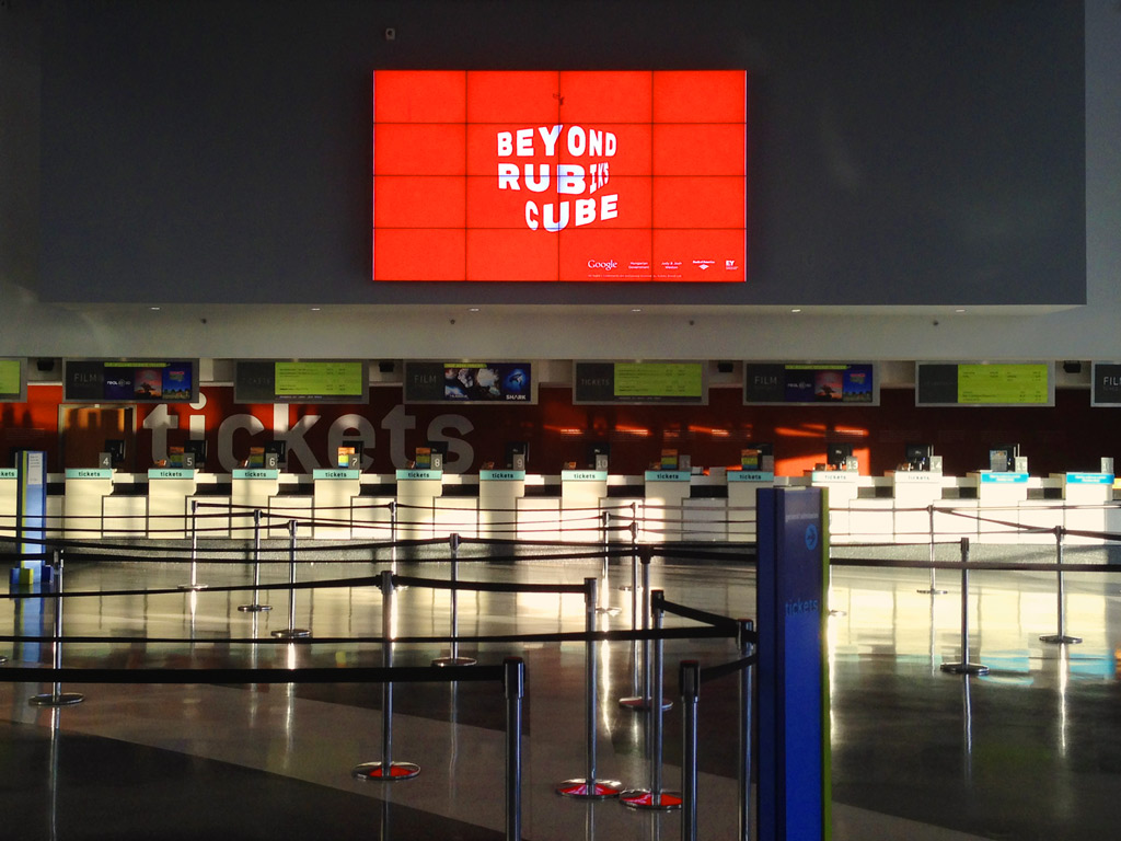 An empty Liberty Science Center entrance hall with sunset light filtering in through the glass entryway as the “Beyond Rubik’s Cube” branding animates above the vacated ticket counters, 2014.