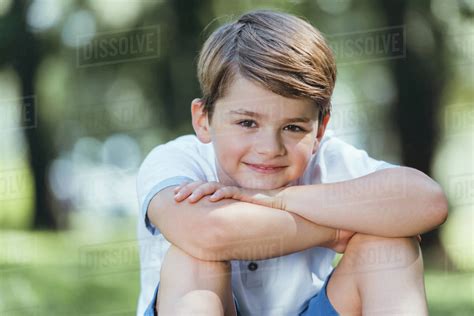 Portrait of cute happy little boy sitting and smiling at camera in park ...