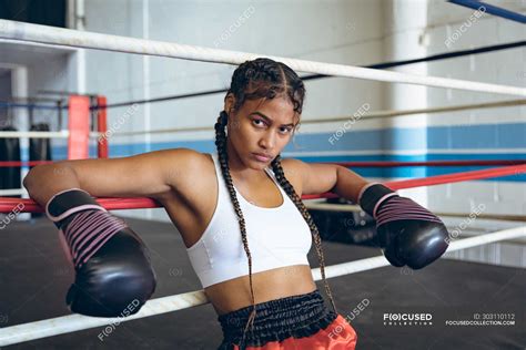 Female boxer with boxing gloves leaning on ropes and looking at camera ...