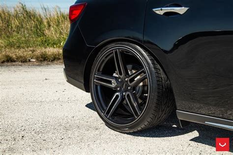 the rear end of a black car parked in a parking lot next to tall grass