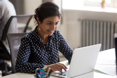 Smiling Indian female employee using laptop at workplace - Helpside