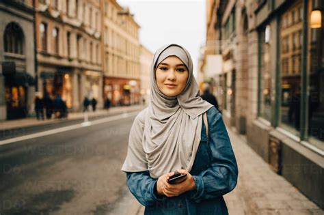 Portrait of confident young woman wearing hijab standing with mobile ...