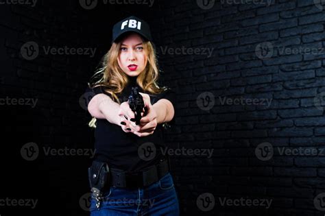 FBI female agent in cap and with gun at studio against dark brick wall ...