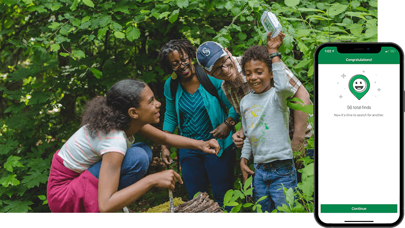 Two adults and two children stand in the forest looking at the youngest who holds the geocache container in the air above his head. 